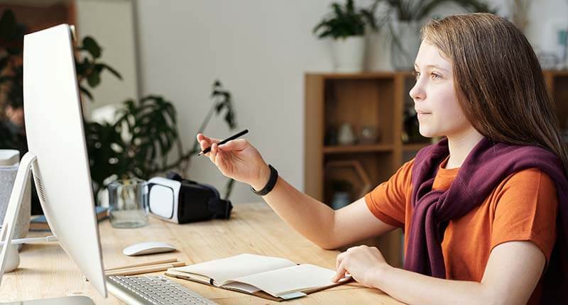 Young girl studying and writing on paper while being tutored over an online meeting